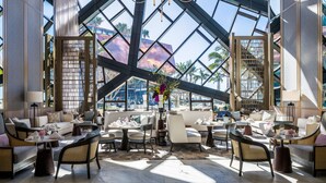 A spacious lounge area with multiple seating options, including sofas and chairs, arranged around a central table. Large windows provide a view of palm trees and a building in the background. - The Langham, Gold Coast and Jewel Residences (Surfers Paradise)
