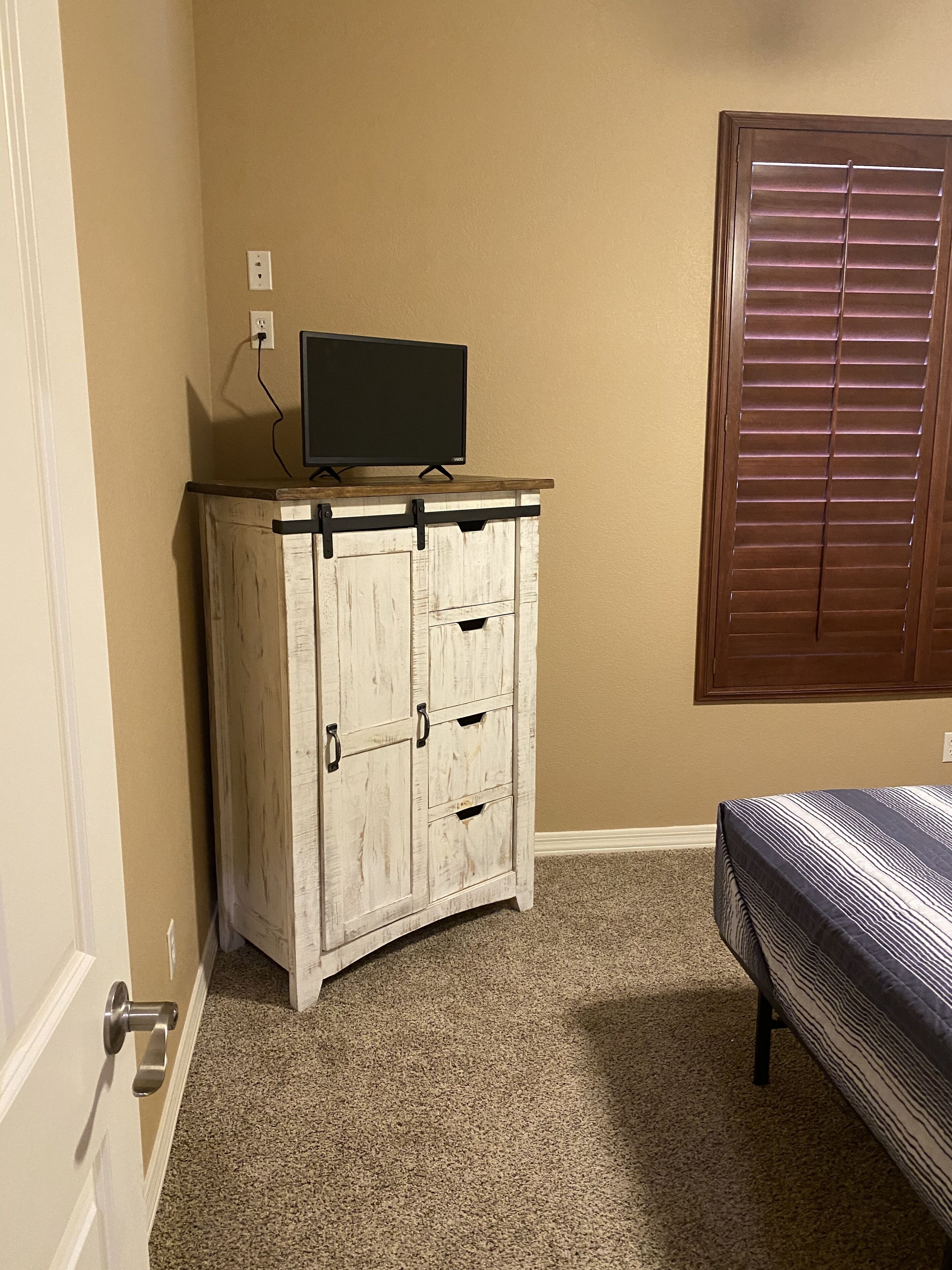 A bedroom with a wooden dresser, a television on top, a window with blinds, and a bed with a striped bedspread.