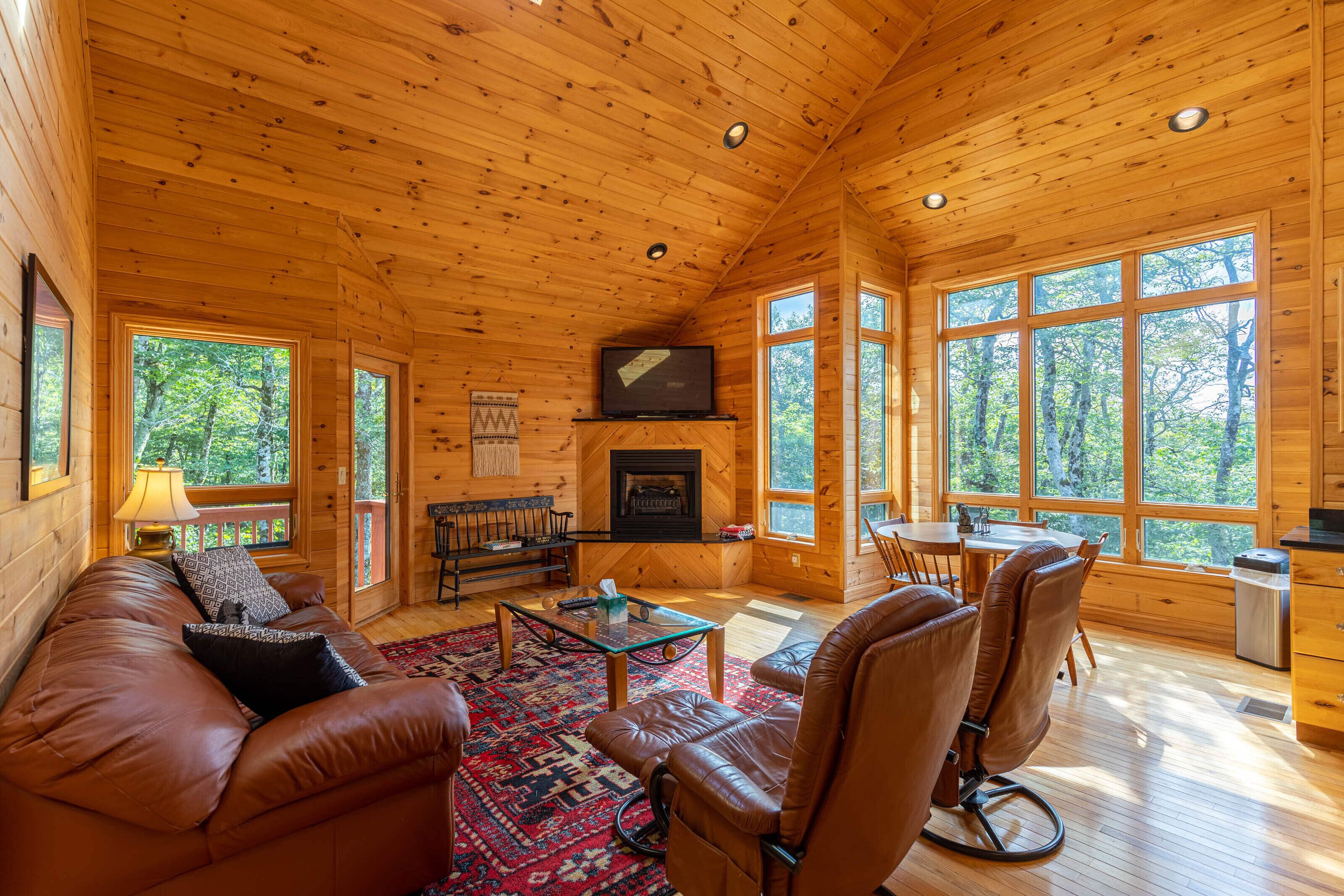 Interior acogedor de una cabaña con techo de madera, chimenea, televisor, sofá, una mesa de centro y ventanas amplias con vistas al bosque.