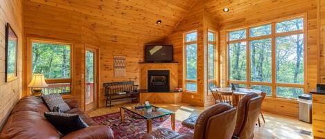 Interior acogedor de una cabaña con techo de madera, chimenea, televisor, sofá, una mesa de centro y ventanas amplias con vistas al bosque.