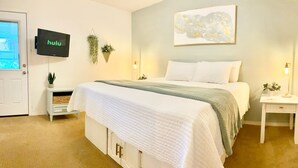 Un dormitorio ordenado con cama, televisión montada en la pared, mesita de noche con lámpara y una planta pequeña.