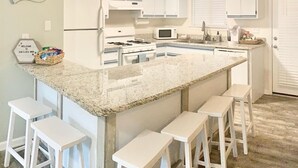 Una cocina con armarios blancos, encimera de granito, estufa a gas, microondas y taburetes de bar.