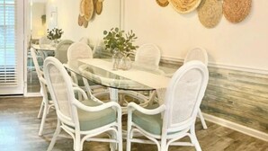 Un comedor con una mesa de vidrio rodeada de sillas blancas, un florero con plantas y adornos de pared tejidos.