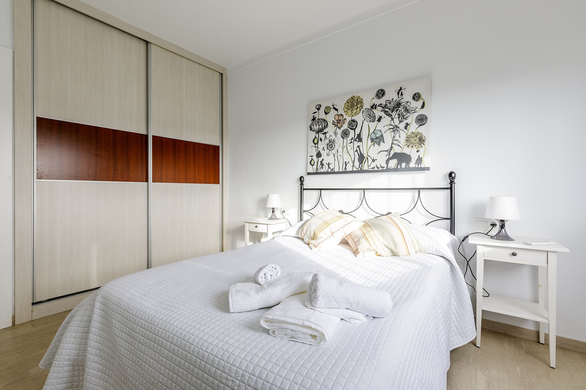 A neatly made bed with white bedding and two folded towels, a bedside table with a lamp, and a wall-mounted artwork depicting flowers and animals.