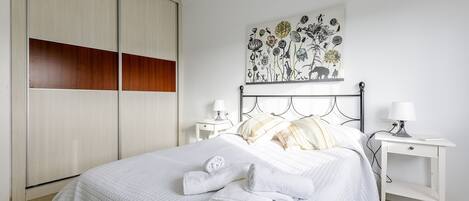 A neatly made bed with white bedding and two folded towels, a bedside table with a lamp, and a wall-mounted artwork depicting flowers and animals.