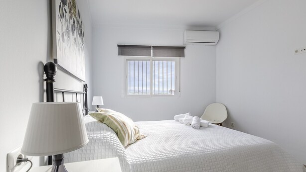 A bedroom with a bed, a bedside table with a lamp, a window with blinds, and an air conditioning unit. - A&n Torrox Beach    . (Torrox Costa)