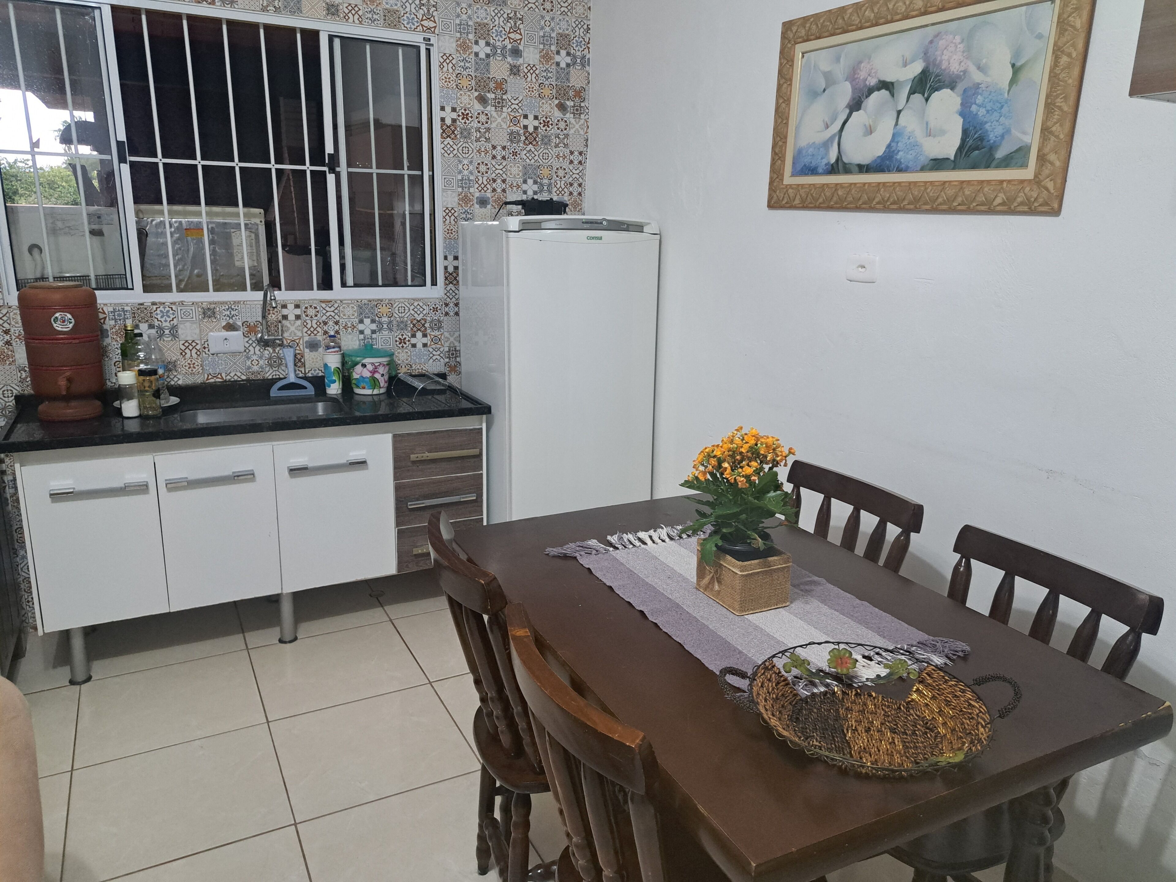 A kitchen with a dining table, chairs, a refrigerator, and a window with bars.