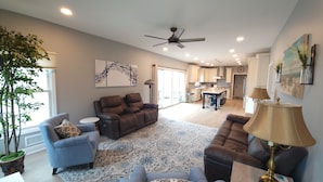 A spacious living room with a large sectional sofa, a blue armchair, and a coffee table. There is a kitchen in the background with white cabinets and a dining area. - 5BR/3BTH waterfront family beach house with hot tub, kayaks, bikes and game room (Mantoloking)