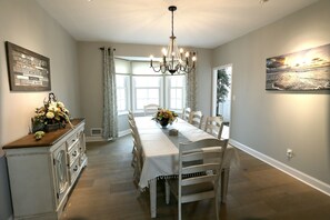 Une salle à manger avec une longue table, des chaises blanches, un lustre et une fenêtre avec des rideaux.