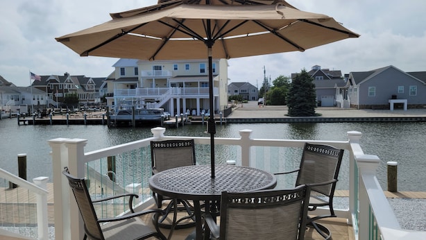 A deck with a table and chairs, overlooking a marina with boats and waterfront buildings. - 5BR/3BTH waterfront family beach house with hot tub, kayaks, bikes and game room (Mantoloking)