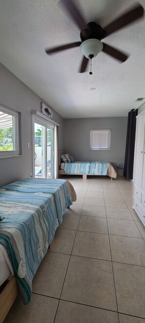 Quarto com cama, ventilador de teto, janela e porta que dá para área externa.