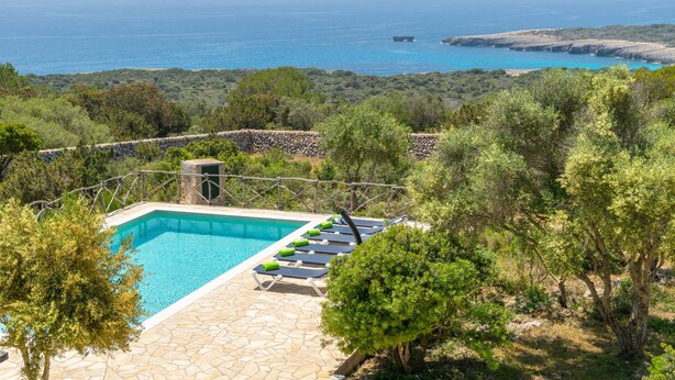 Piscina con sillones de descanso, rodeada de baldosas de piedra y vegetación, con vistas a un paisaje costero.