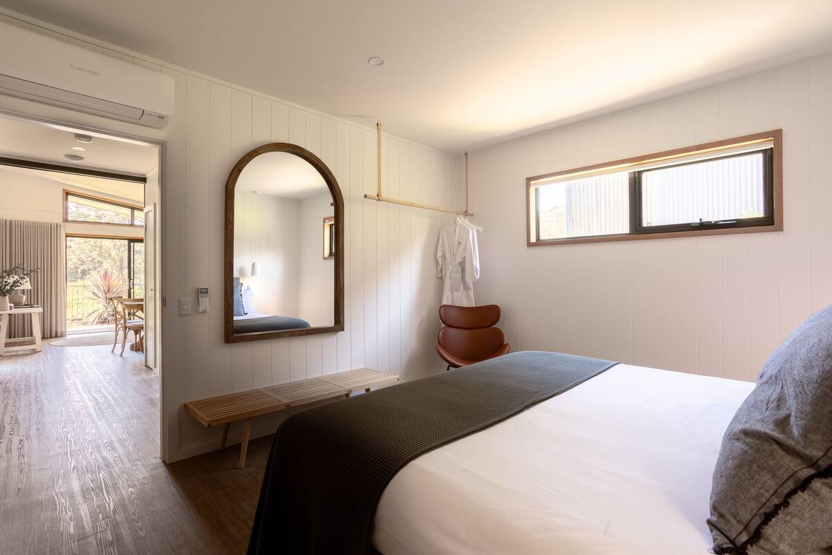 A bedroom with a large bed, a wooden bench, a mirror, a window, and a view of an outdoor area.