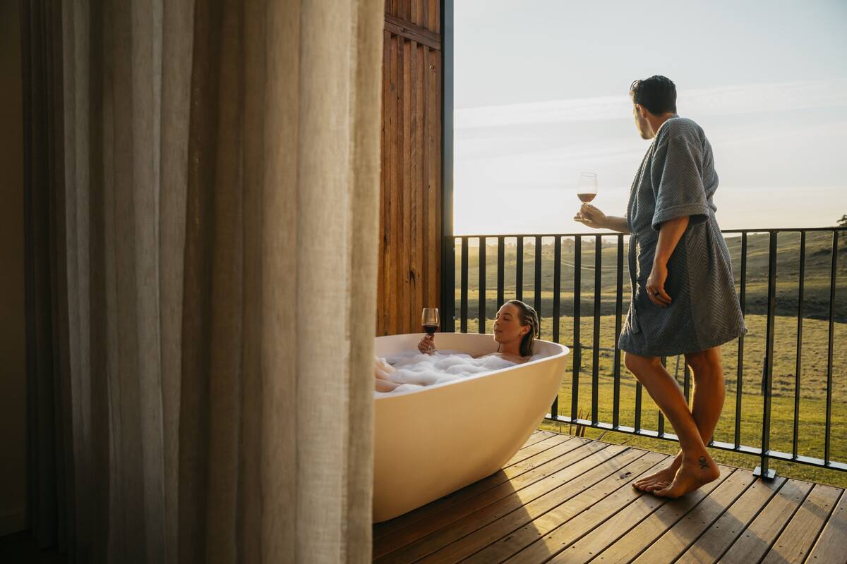 A person standing on a wooden deck holding a glass of wine, overlooking a large bathtub filled with bubbles.