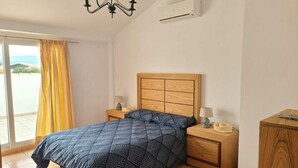 A bedroom with a bed, bedside tables, a nightstand, a wooden headboard, an air conditioning unit, and a view through a window. - Apartment ANDRES in the center of Els Poblets (Poblets (els))