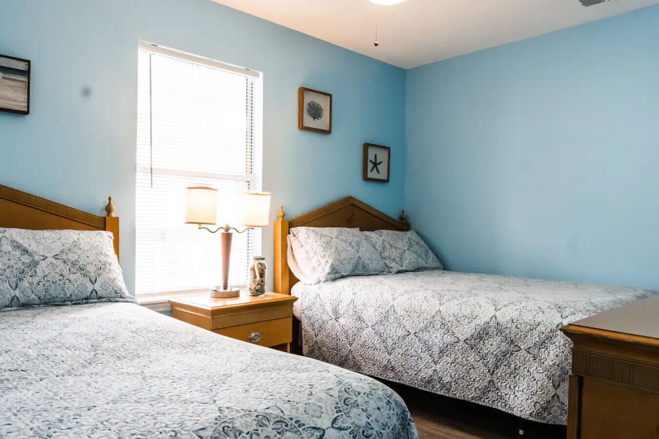 A bedroom with two beds, a window with blinds, a bedside table with a lamp, and a clock on the wall.
