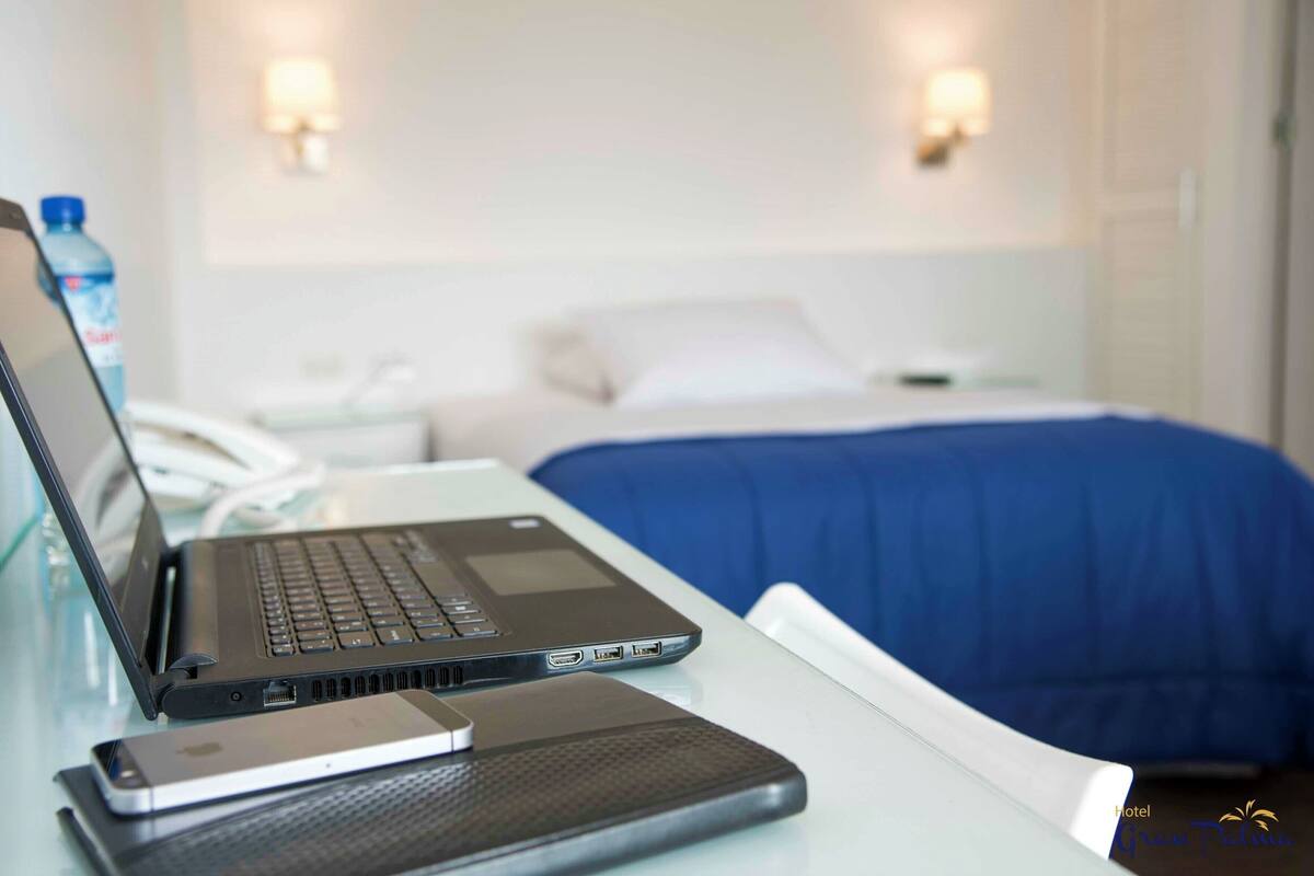 Una laptop sobre una mesa con un teléfono inteligente a su lado, una cama con una manta azul y una botella de agua al fondo.