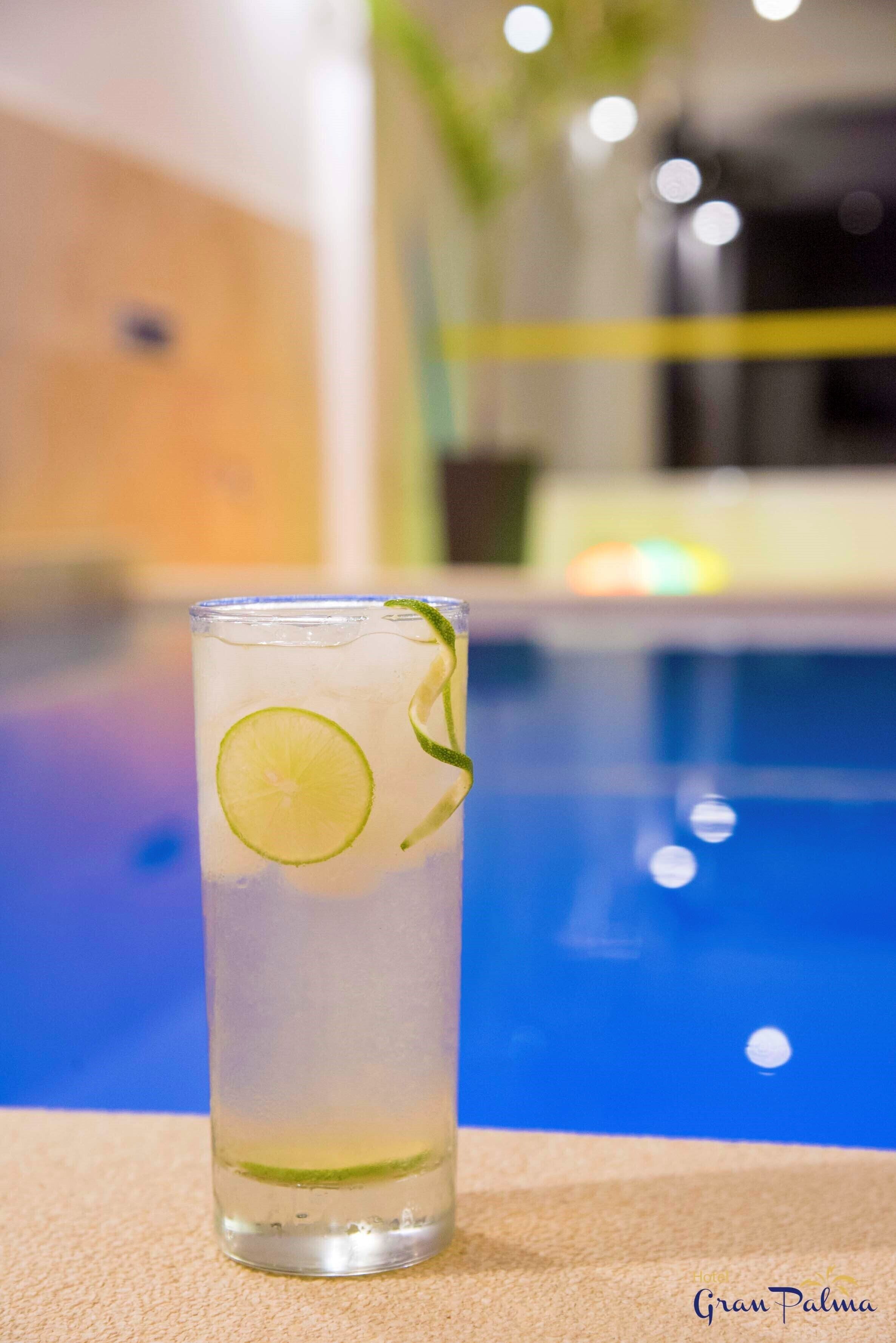 Un vaso alto con una rodaja de lima y una pajita verde, sobre una superficie color beige cerca de una piscina.