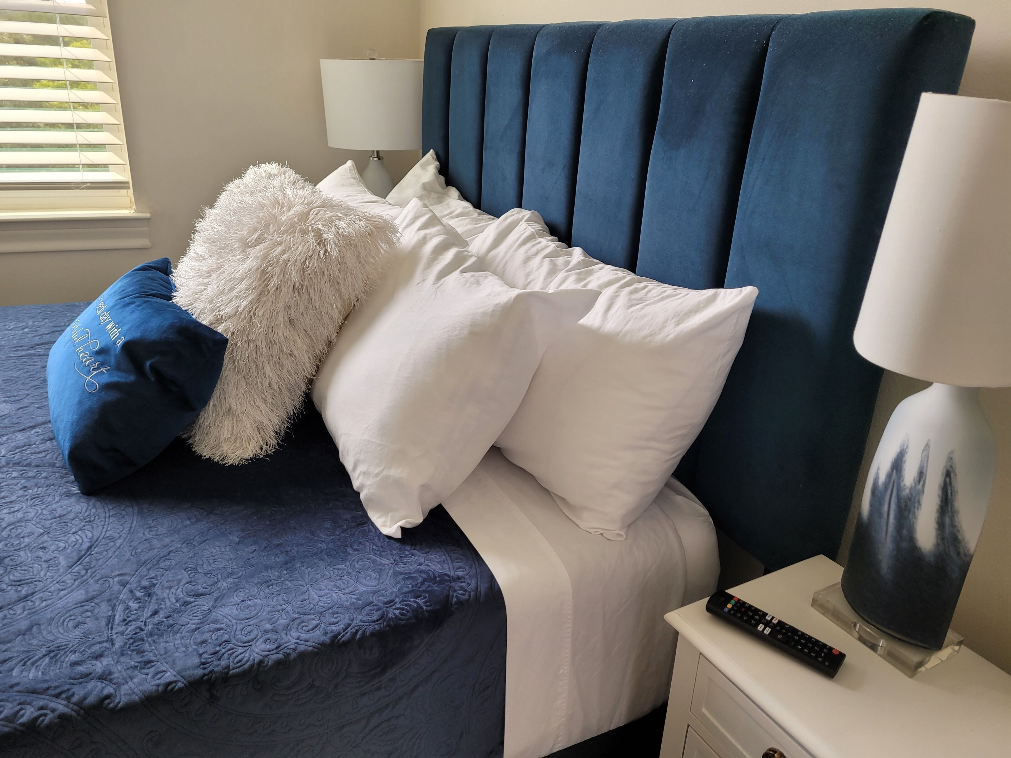 A neatly made bed with a blue headboard, white pillows, and a fluffy white pillow. A remote control and a lamp are on a bedside table.