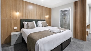 A modern hotel room with a large bed, wooden panel walls, a glass shower, and a wooden partition.