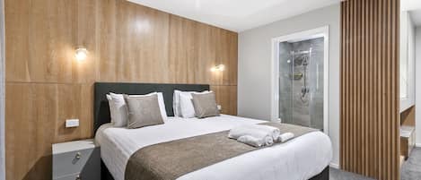 A modern hotel room with a large bed, wooden panel walls, a glass shower, and a wooden partition.