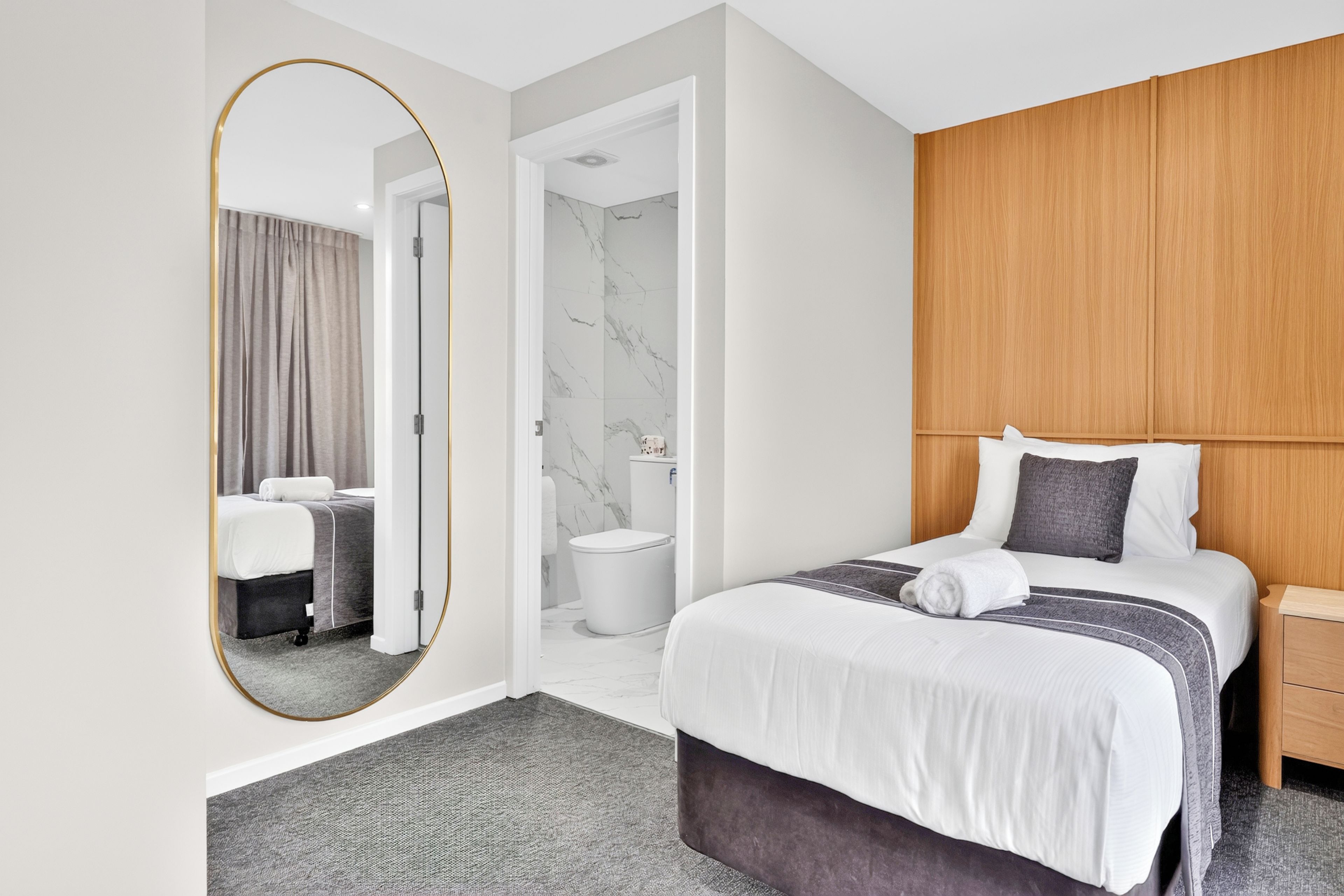 A hotel room with a large mirror, a bed with a grey and white striped bedspread, a wooden headboard, a bedside table, and a grey carpet.