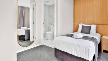 Une chambre d’hôtel avec un grand miroir, un lit recouvert d’une couvre-lit à rayures grises et blanches, une tête de lit en bois, une table de chevet et un tapis gris.