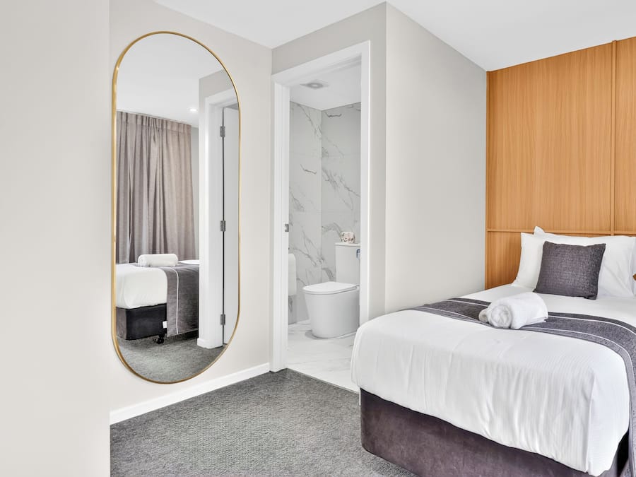 Une chambre d’hôtel avec un grand miroir, un lit recouvert d’une couvre-lit à rayures grises et blanches, une tête de lit en bois, une table de chevet et un tapis gris.