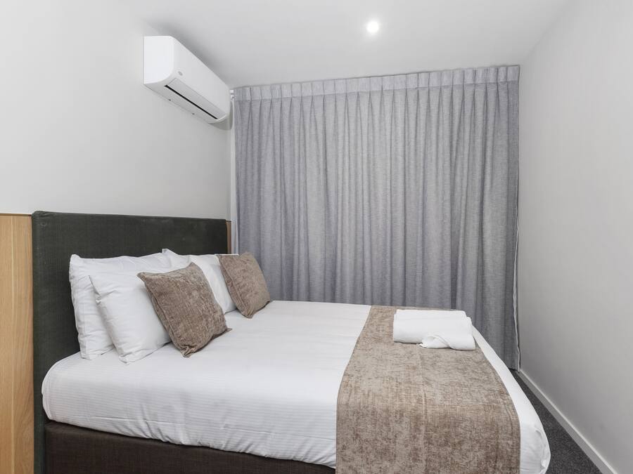 A neatly made bed with white linens and a brown throw blanket, a dark headboard, and a grey curtain.