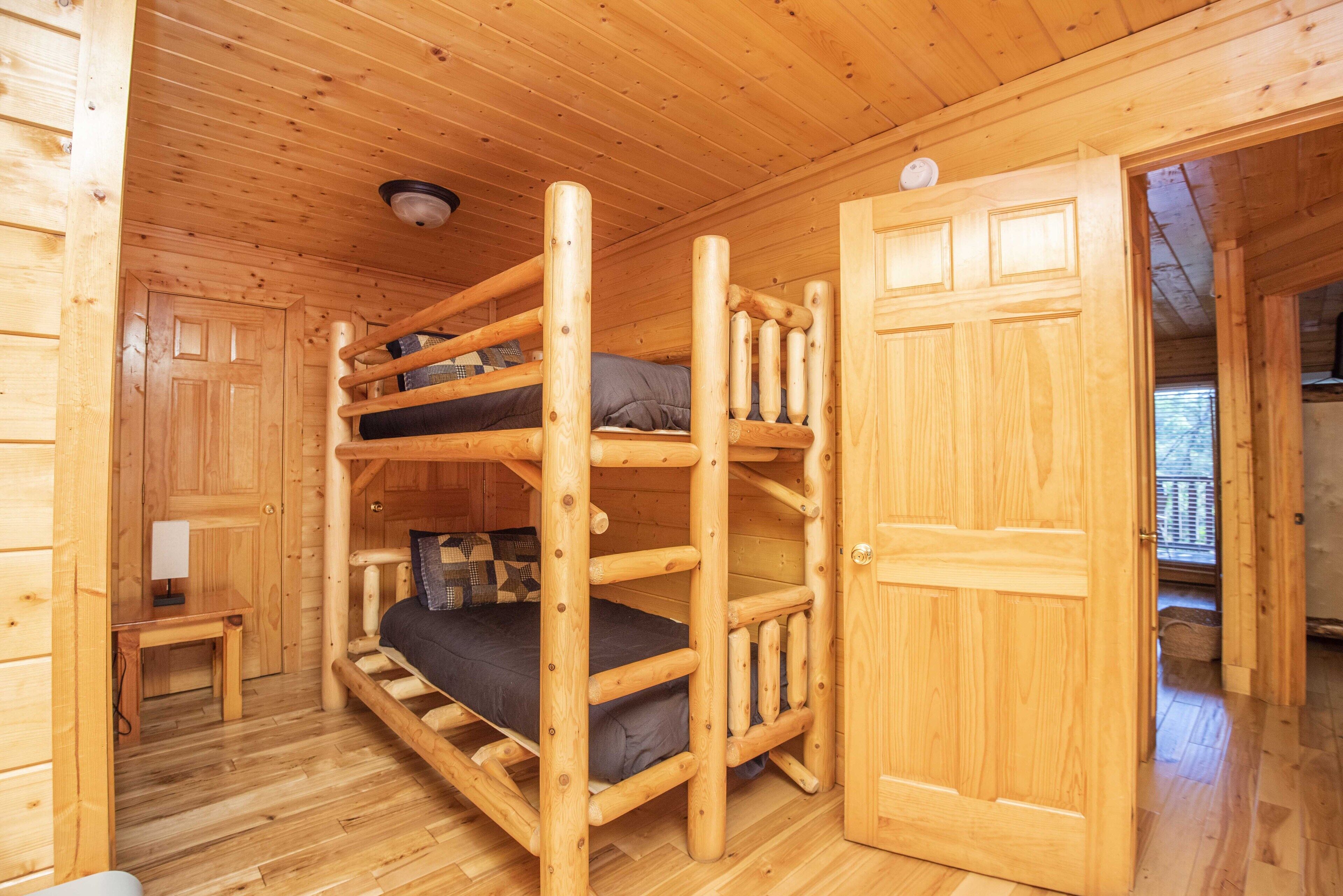 A wooden cabin with a bunk bed, a small table with a lamp, and a door leading to another room.