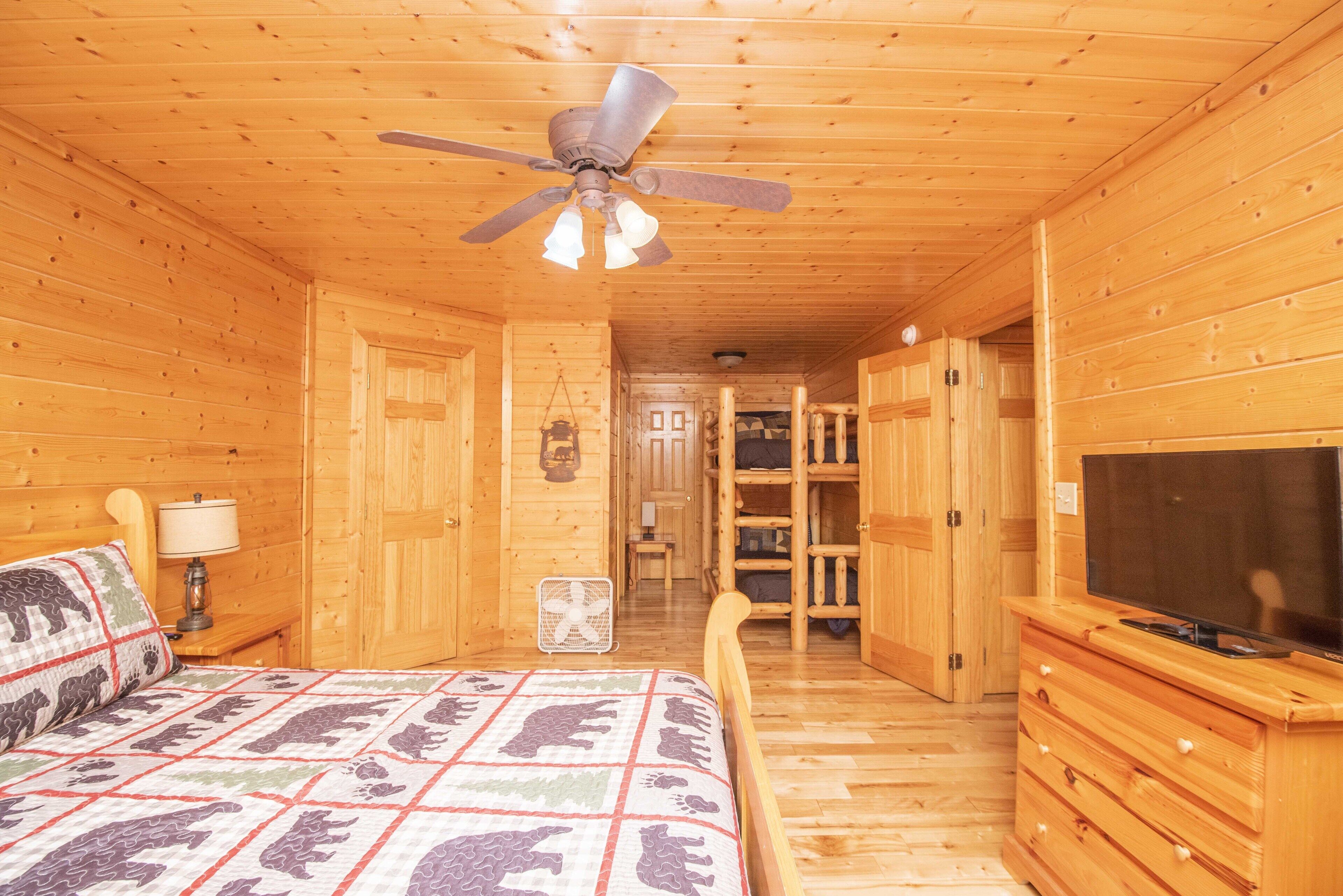 A wooden cabin room with a bed, a ceiling fan, a TV, and a small fan.