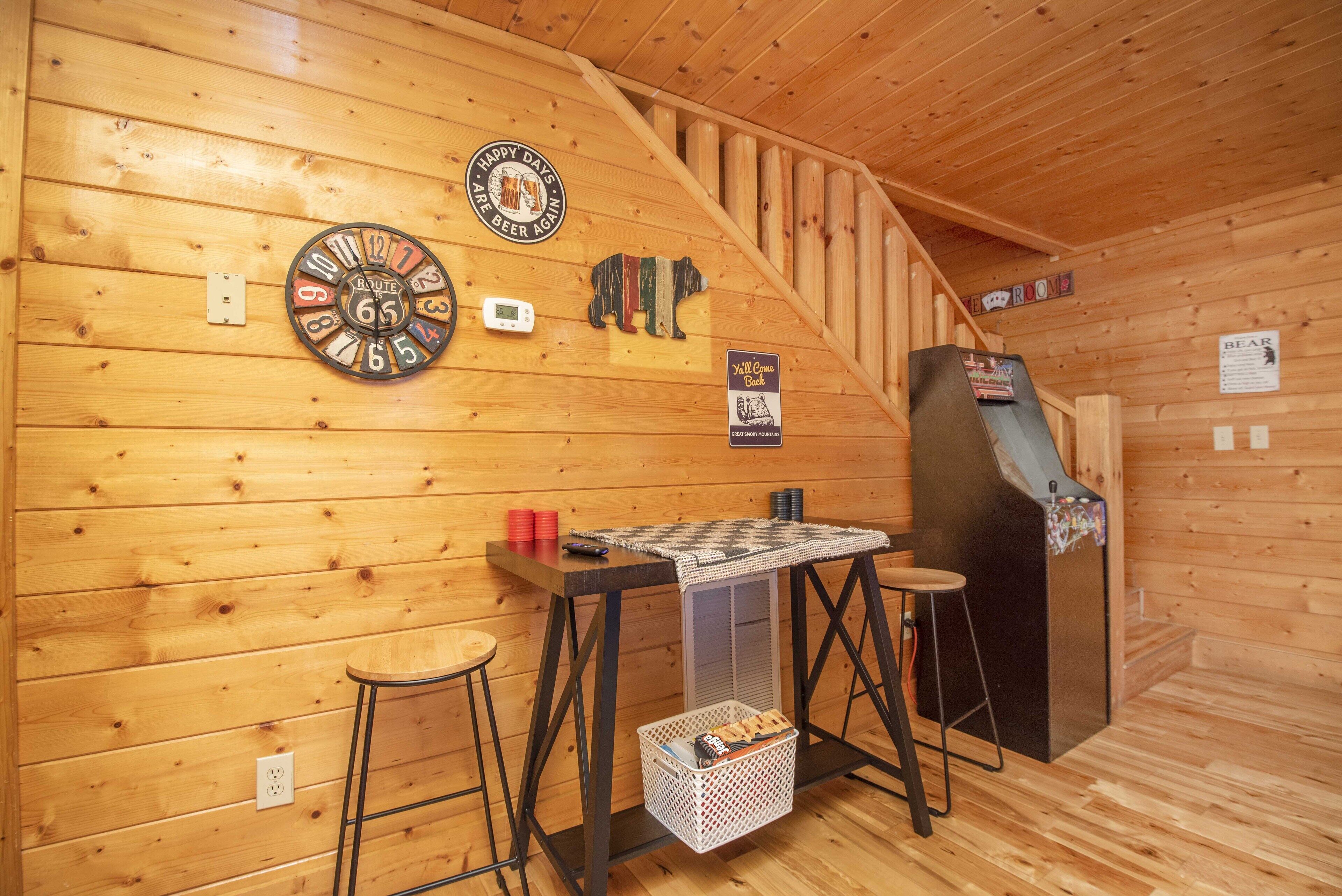 Interior de una cabaña de madera con escalera de madera, dardos, un reloj y una pequeña mesa con taburetes.