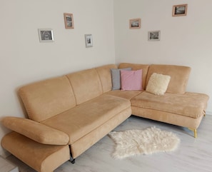 A tan sectional sofa with a fluffy white rug, a light-colored wooden floor, and several framed pictures on the wall.