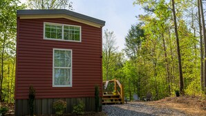A red cabin with two windows, a wooden deck, and a gravel path leading to a chair and table. - Delightful Forest Tiny Home with Hot Tub (Murphy)