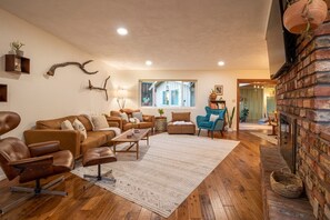 A cozy living room with a brown leather sofa, a blue armchair, and a wooden coffee table. A large window provides natural light, and a brick fireplace adds warmth.
