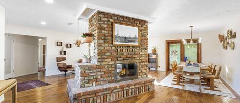 A spacious living room with a brick fireplace, wooden flooring, and a dining area with a table and chairs.