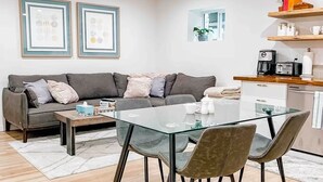 A modern living room with a grey sofa, a glass dining table, and a kitchen area with a coffee machine and shelves. - 3 bdrm suite w/ rooftop patio, firetable & sauna + hot tub, cold plunge & more! (Kelowna)