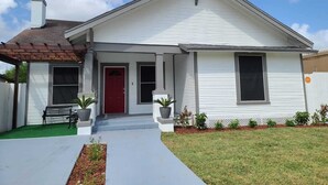 A single-story house with a red front door, a covered porch, and a small front yard with a bench and potted plants. - Beautiful 3BR&2BA House located in the Historic part of Brownville-SPI-SpaceX-MX (Brownsville)