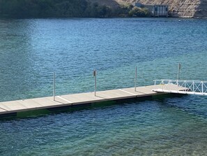 Eine Betonsteg erstreckt sich in einen ruhigen See mit klarem Wasser, umgeben von einer naturbelagten Landschaft.