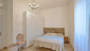 Une chambre avec une tête de lit en bois, un lit recouvert d’une couverture à carreaux, une chaise avec un coussin à motifs floraux et une petite table ronde.