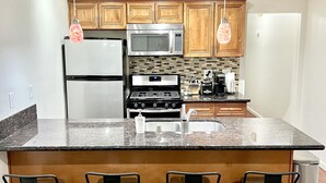 A modern kitchen with wooden cabinets, stainless steel appliances, a granite countertop, and three bar stools. - Resort Style Condo near Disneyland, Beaches, Los Angeles & so much more  (Fountain Valley)