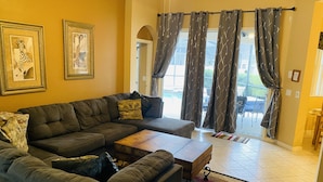 A living room with a sectional sofa, a wooden coffee table, and a patterned rug. There are two framed artworks on the wall and a view of the outdoors through a glass door.