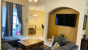 A living room with a sofa, a coffee table, a TV mounted on an arched wall, a dining area with a table and chairs, and a ceiling fan.