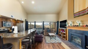 A spacious living area with a kitchen, bar stools, a fireplace, and a large window overlooking a mountainous landscape. - New Lamartine Condos Building 2 Unit #2533 by Summit County Mountain Retreats (Silverthorne)