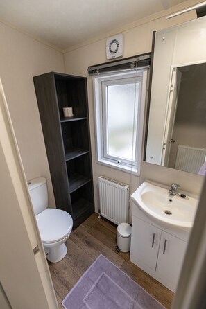 A compact bathroom with a toilet, a white sink, a mirror, a window, and a dark wooden shelf. - The Retreat, Sandhills Holiday Park by the beach in Mudeford (Christchurch)