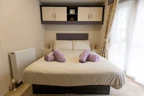 A neatly made bed with two rolled towels, a bedside table with a lamp, and a window with curtains. - The Retreat, Sandhills Holiday Park by the beach in Mudeford (Christchurch)