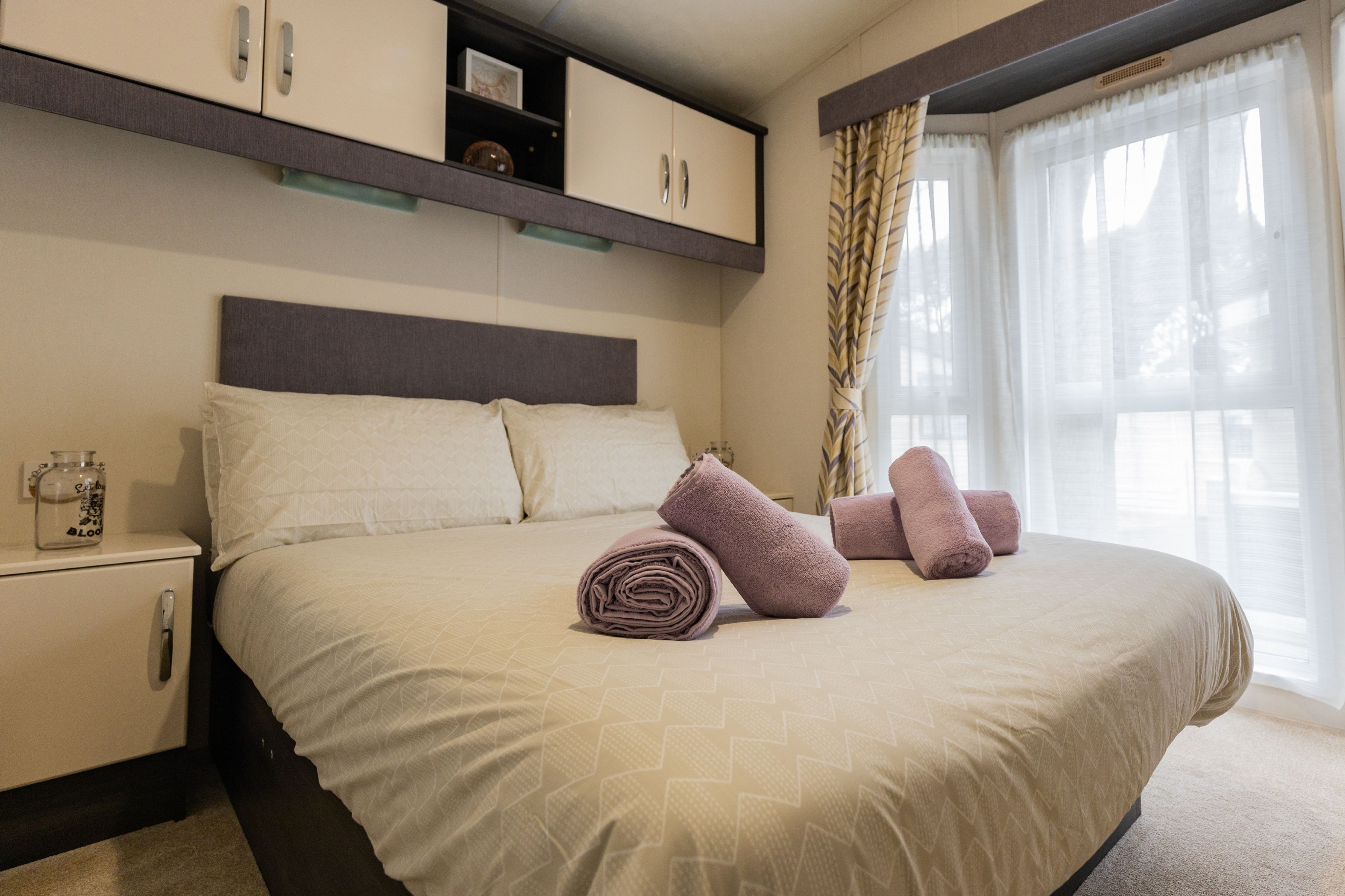 A neatly made bed with rolled towels, a bedside table with a glass container, and a built-in wardrobe above the bed.