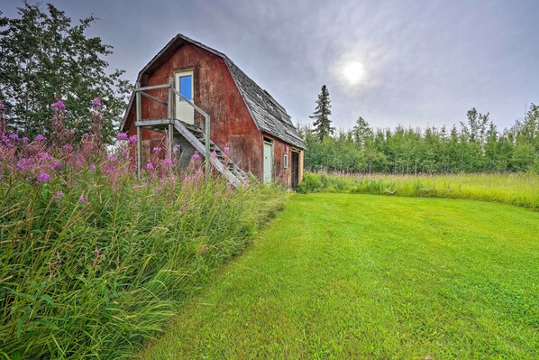 A rustic barn with a wooden staircase leading to a blue door, surrounded by lush greenery and wildflowers.