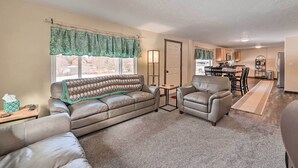 Ein Wohnzimmer mit einer Couch, Sesseln und einem Couchtisch. Im Hintergrund ist eine Küche mit einem Esstisch und Stühlen zu sehen.