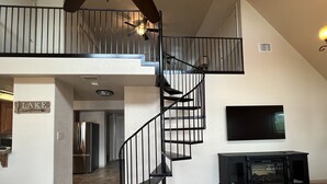 A spacious living area with a black spiral staircase leading to an upper level. A flat-screen TV is mounted on the wall, and a fireplace is visible in the foreground.